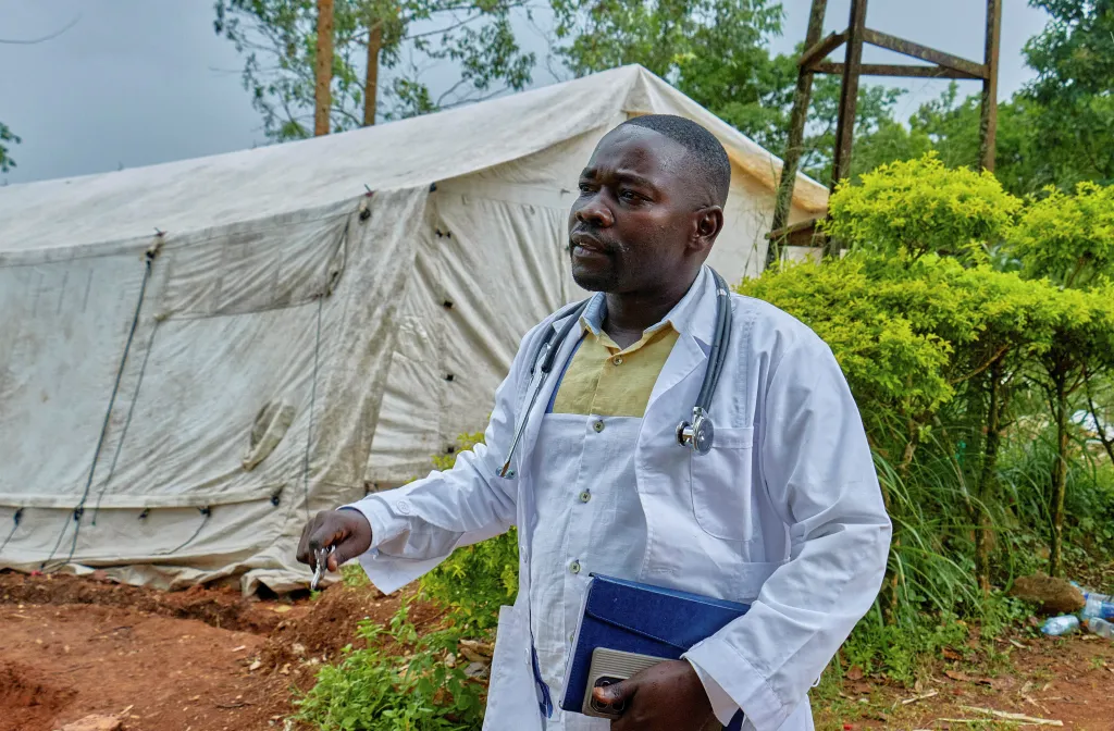 Medical director Richard Lokudu stands in a white lab coat with a stethoscope around his neck, in front of a white tent.
