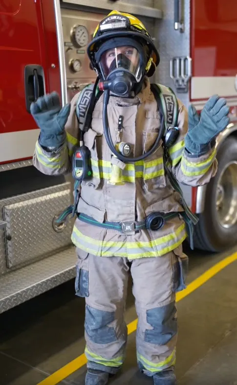 A female firefighter in full gear, including helmet, mask, and breathing apparatus, stands with arms raised.