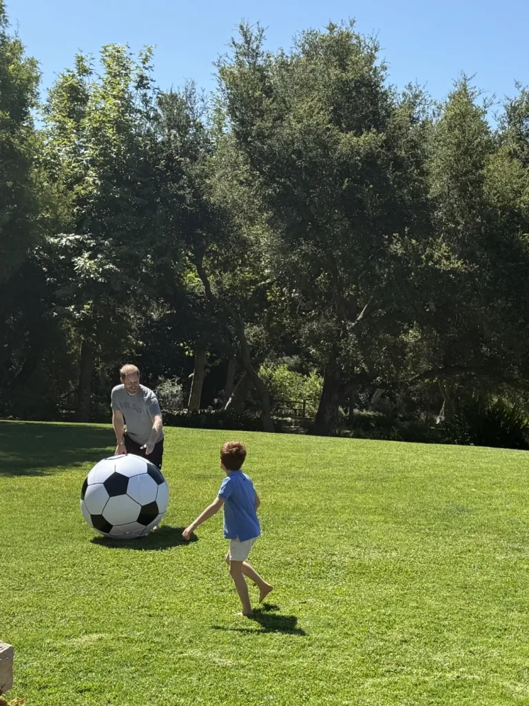 Prince Harry and his son Archie playing with a giant soccer ball in a sunny backyard.