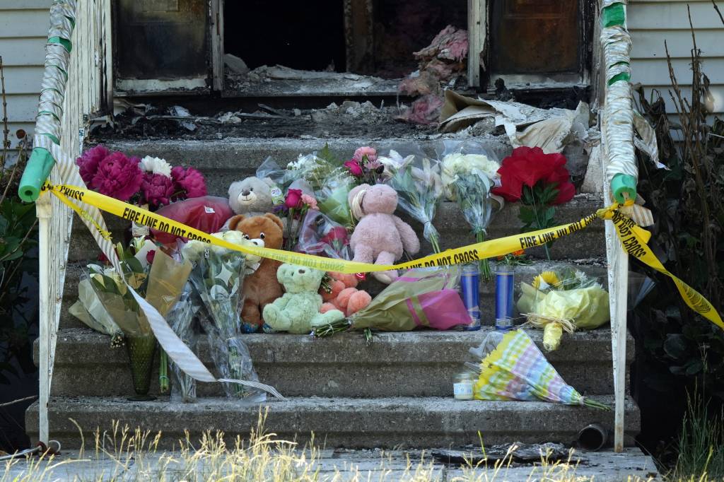A memorial of flowers, candles, and teddy bears on the front steps of a burned-out home, with 