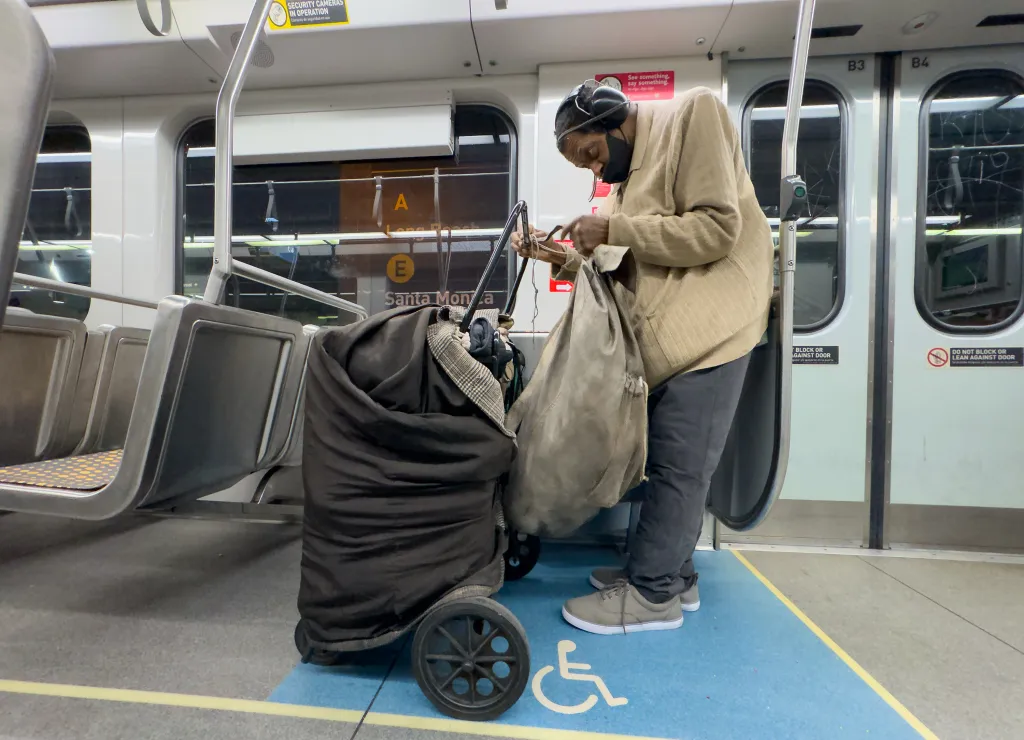 A person on a Los Angeles Metro train adjusts their bag next to a loaded cart in the designated wheelchair area.