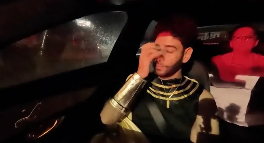 Mido sits in a car, dressed in a black and gold outfit, with his hand on his face while a red figure is next to him.