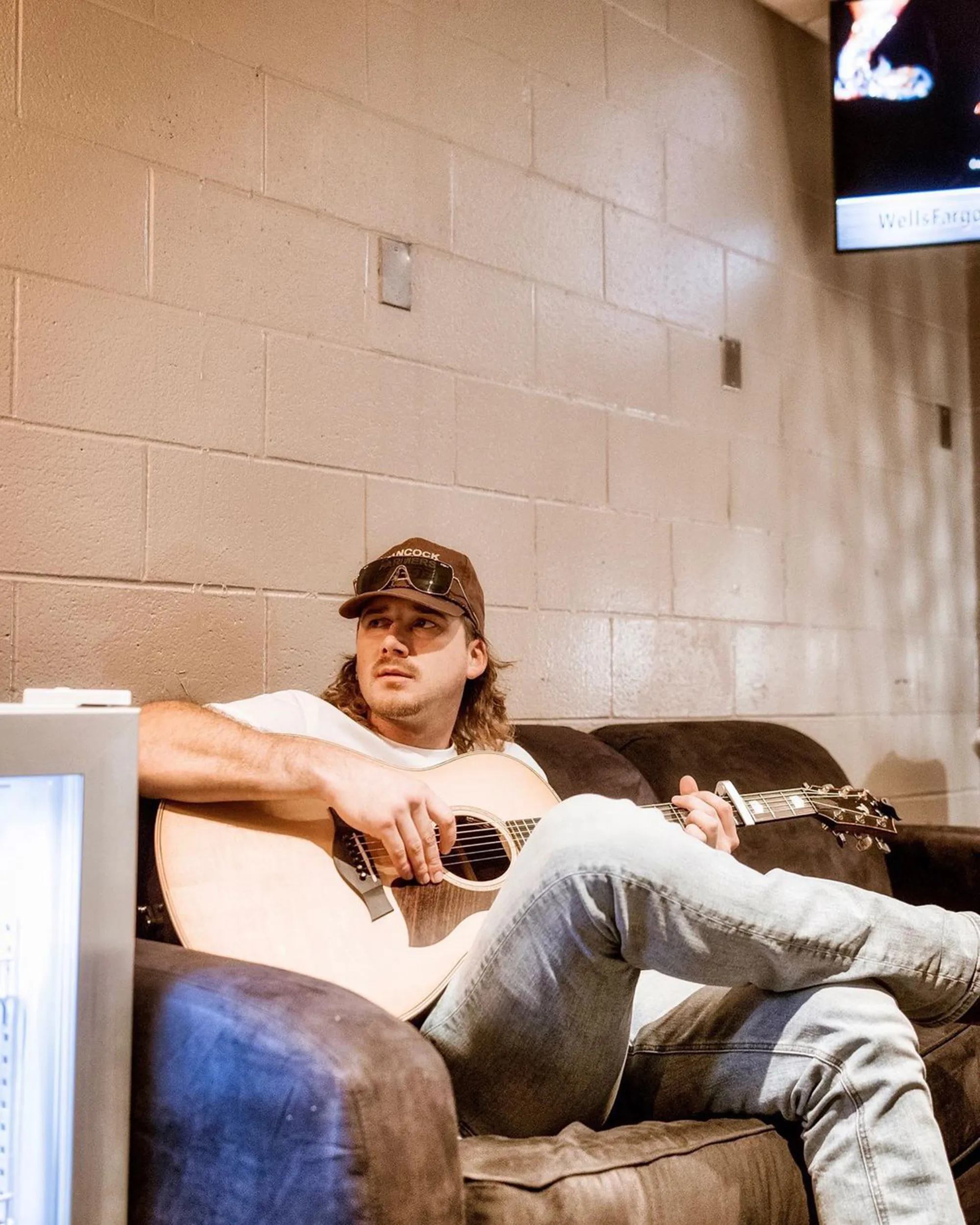 Morgan Wallen sitting on a couch and playing an acoustic guitar.