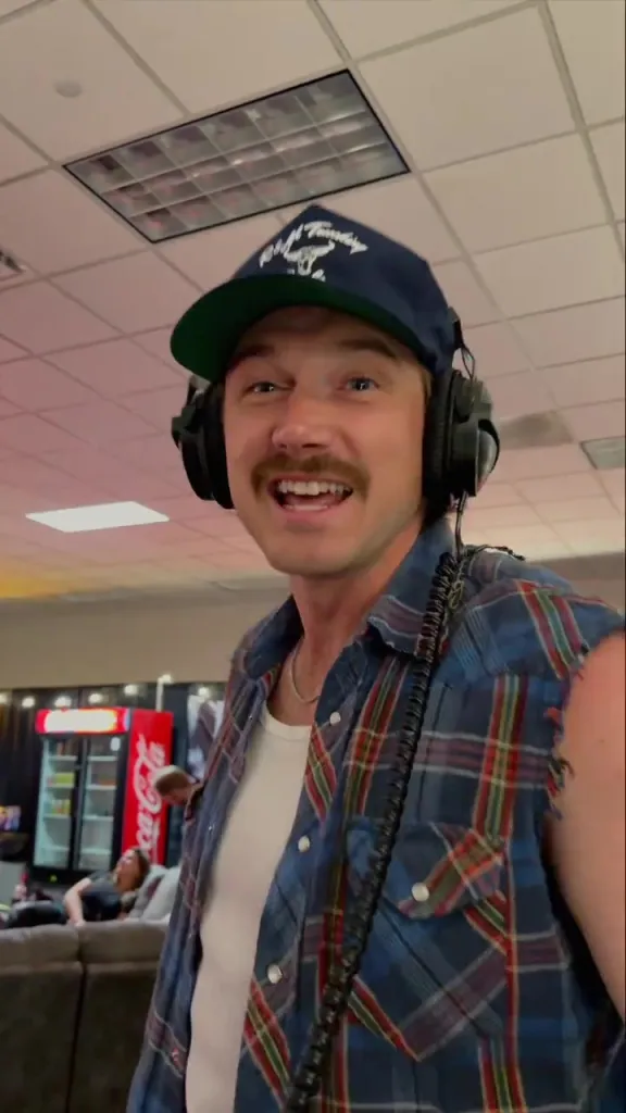 Morgan Wallen smiling and wearing headphones, a blue plaid shirt, and a baseball cap.