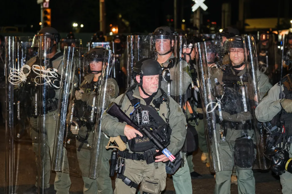 Law enforcement officers in riot gear and helmets stand in a line holding clear shields.
