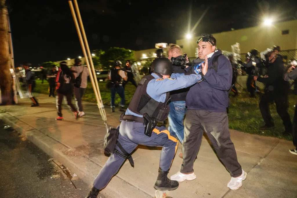 A police officer in riot gear pushing a protester with hands up, while other officers and protesters are visible in the background at night.