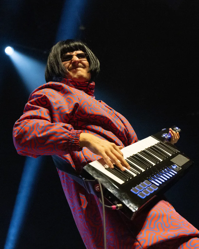Musician Oliver Tree wearing a red and blue patterned jumpsuit, black wig, and sunglasses, playing a keytar on stage.