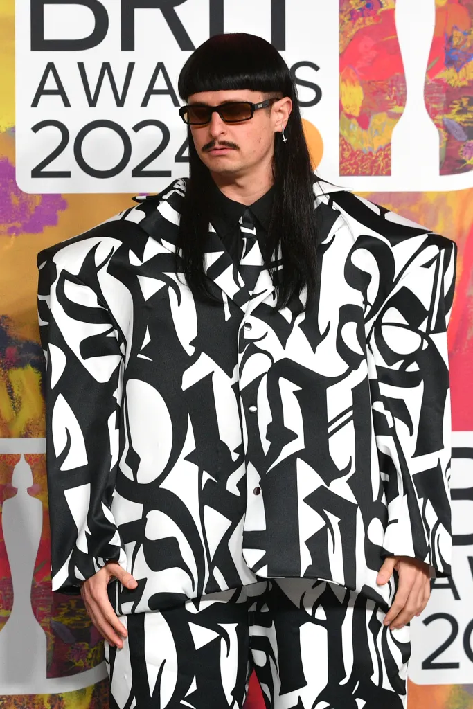 Musician Oliver Tree wearing a black and white patterned suit and sunglasses, standing in front of a BRIT Awards 2024 sign.
