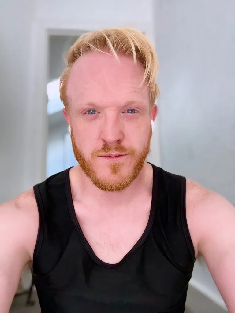 A man with red hair and a beard, wearing a black tank top and earbuds, looks directly at the viewer.