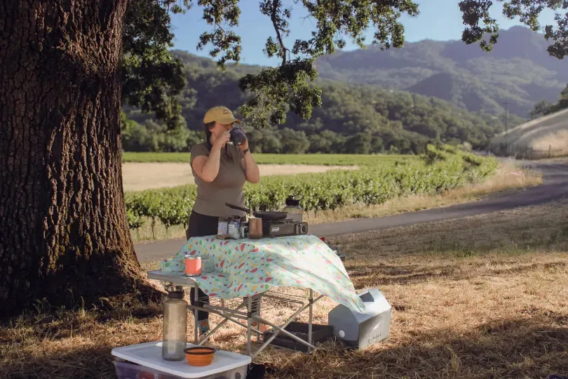 A woman camping next to a vineyard sips from a mug while standing by a portable stove.