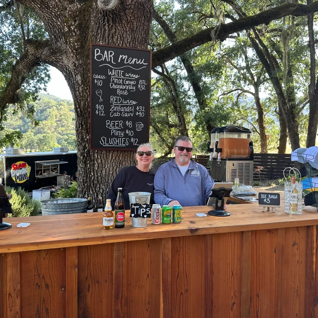 Two people behind a wooden bar under a tree at Nelson Family Vineyards.