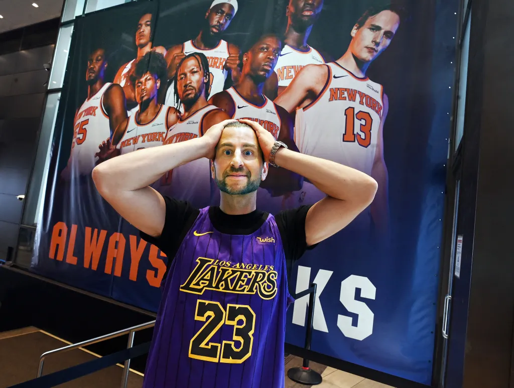Evan Perlmutter, a former Knicks fan, poses in a Lakers jersey with a large Knicks banner behind him.
