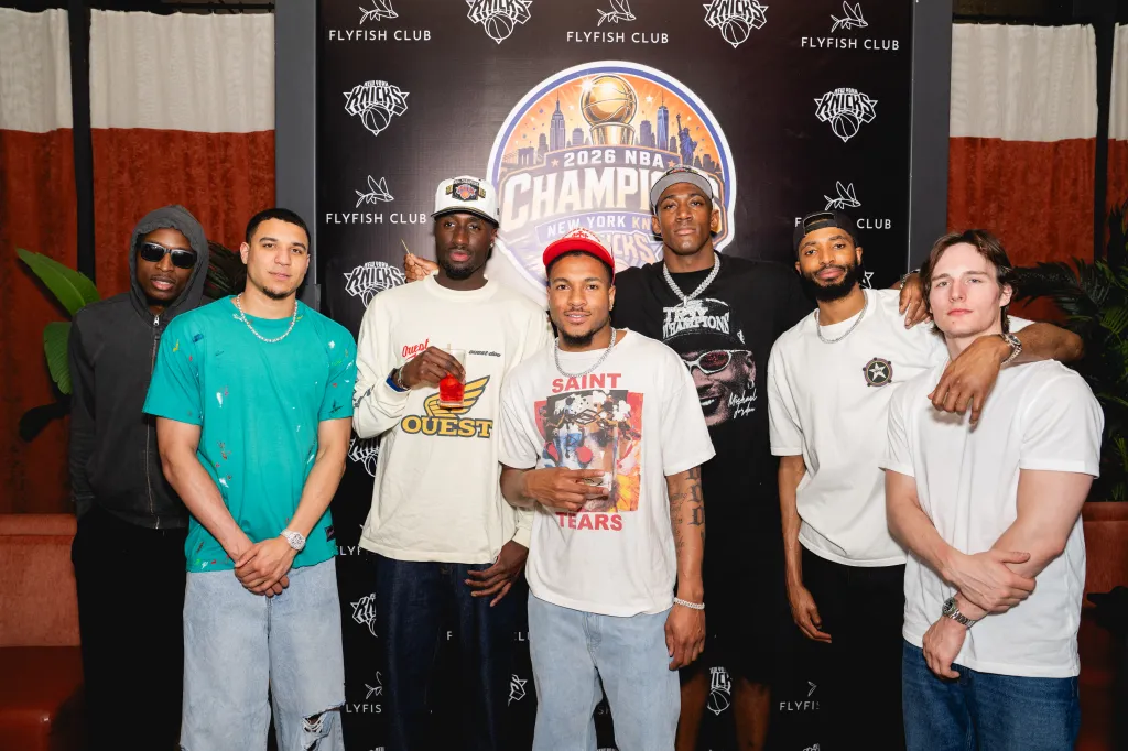 Group photo of six men, including Landry Shamet, Miles McBride, and Josh Hart, standing in front of a banner celebrating the 2026 NBA Championship for the New York Knicks.
