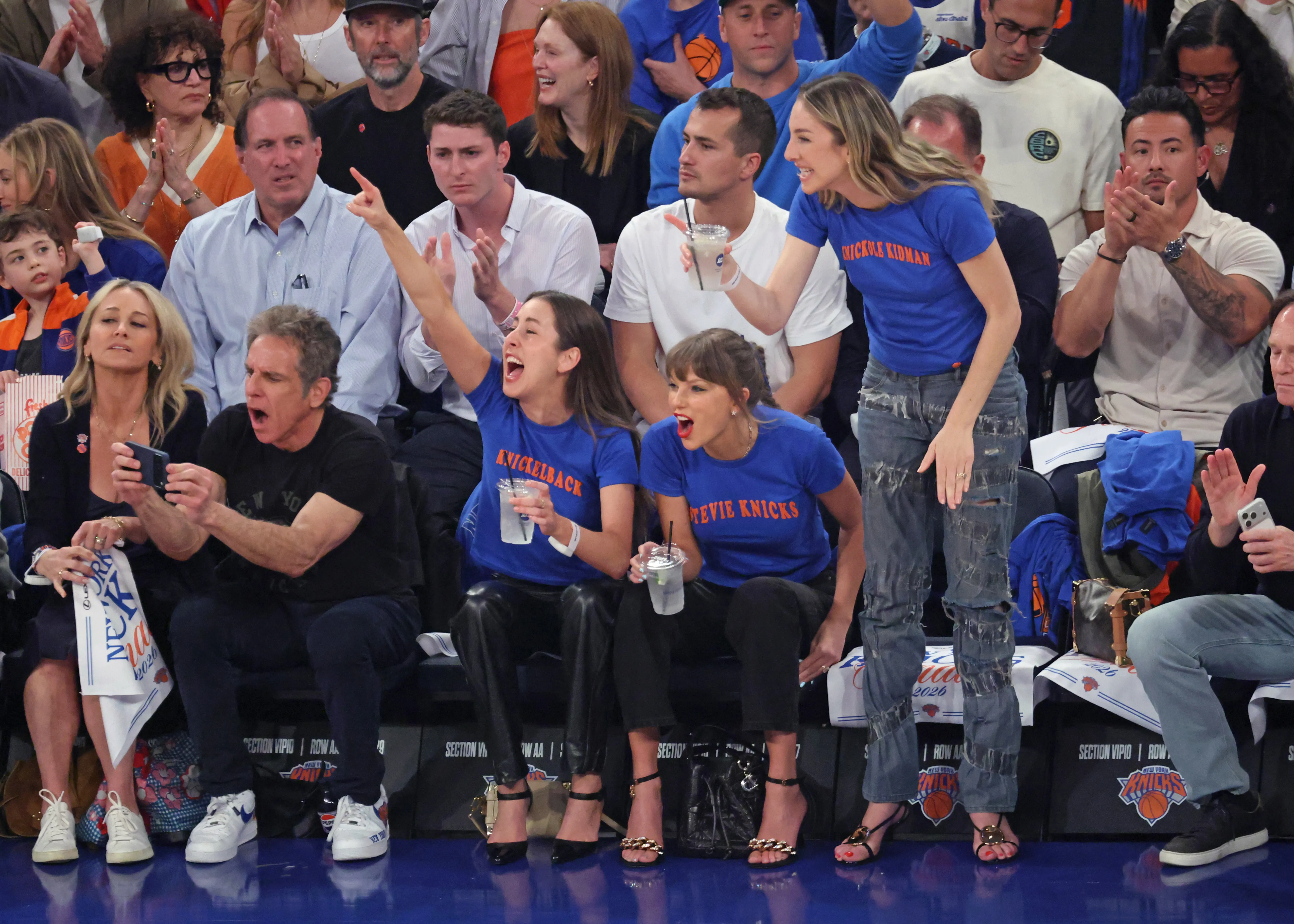 Taylor Swift and the Haim sisters supporting the Knicks at a game, wearing custom t-shirts, with Ben Stiller and Christine Taylor nearby.