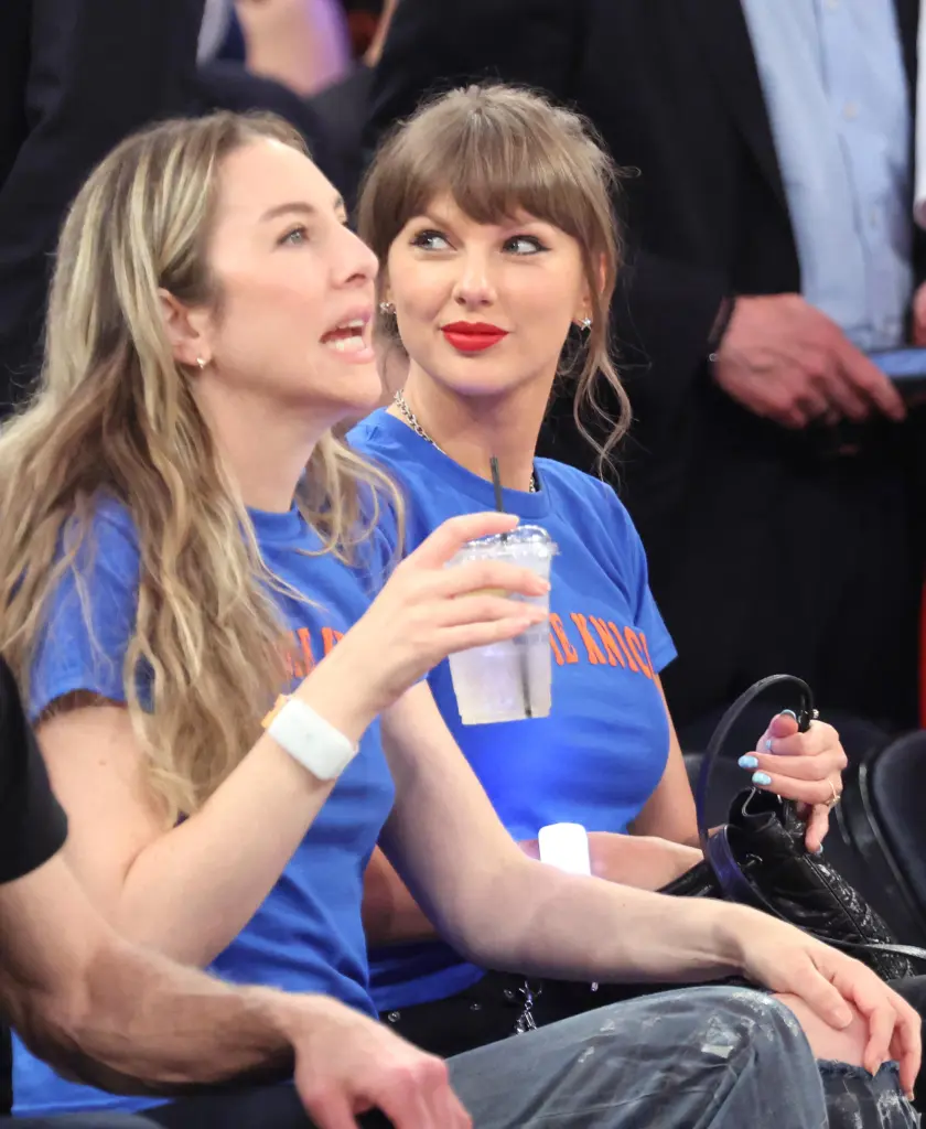 Taylor Swift in a blue t-shirt and red lipstick watching a game with another woman.