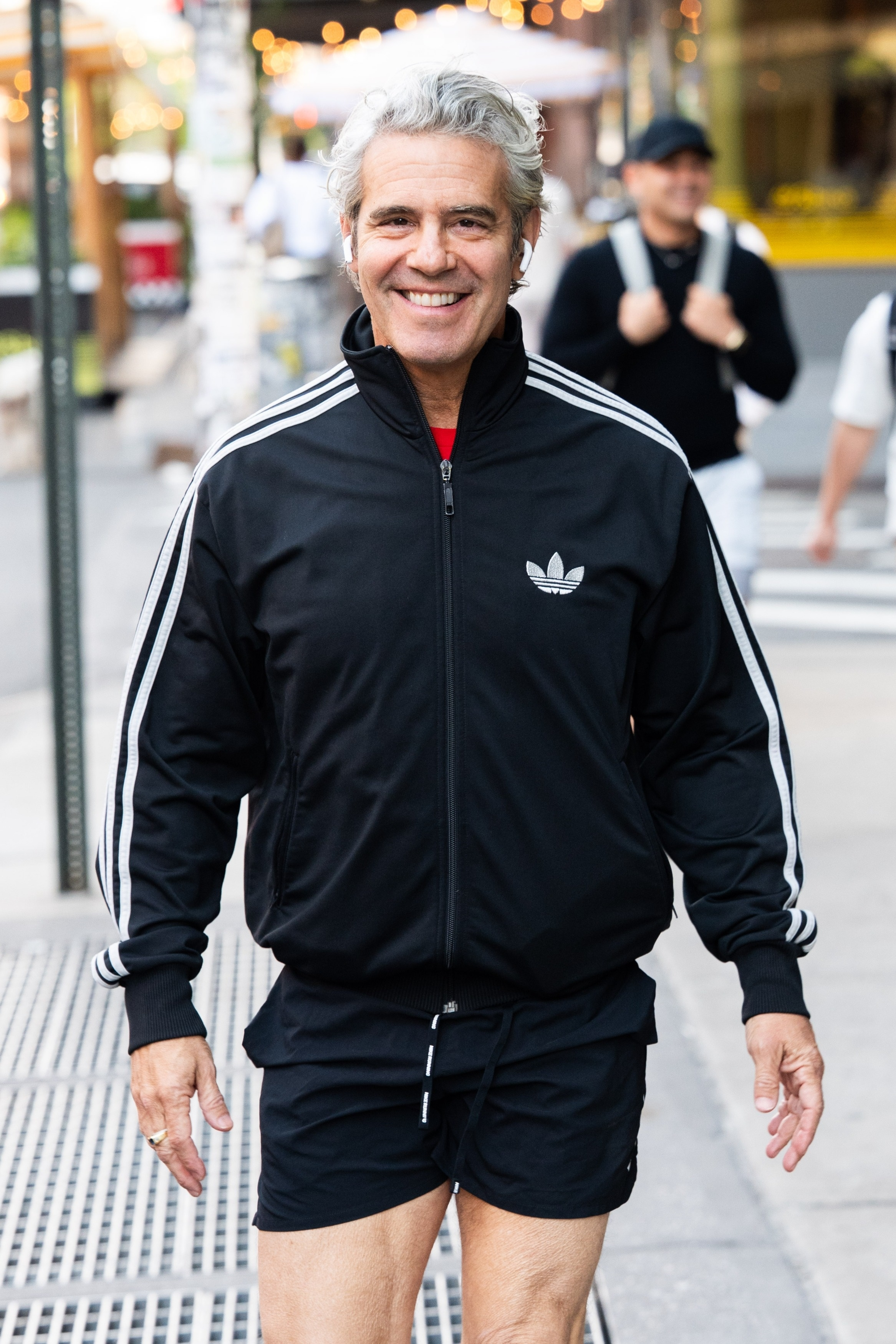 Andy Cohen smiling while wearing a black Adidas track jacket and black running shorts.