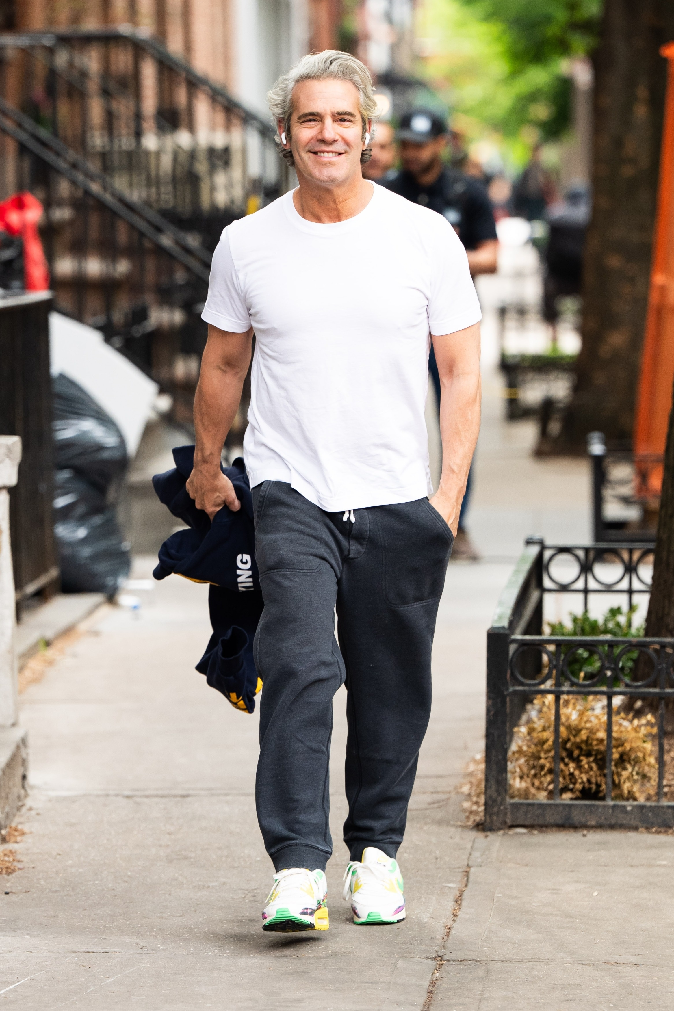 Andy Cohen smiling while walking down a New York street in a white tee, gray sweatpants, and holding a blue hoodie.
