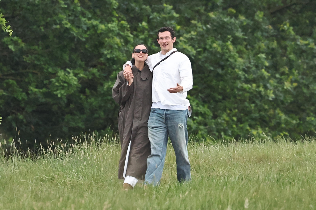 Dua Lipa in a taupe trench coat and Callum Turner in a white shirt and jeans walking through a grassy field.