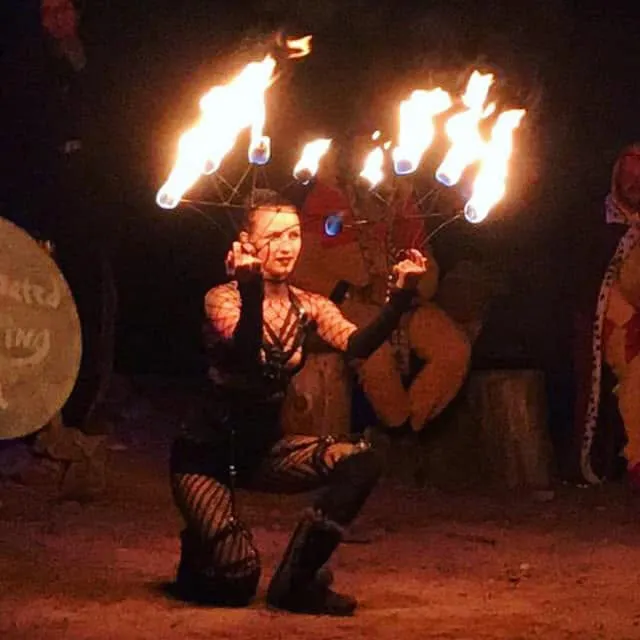 A woman in dark, netted clothing crouches and holds two flaming objects.