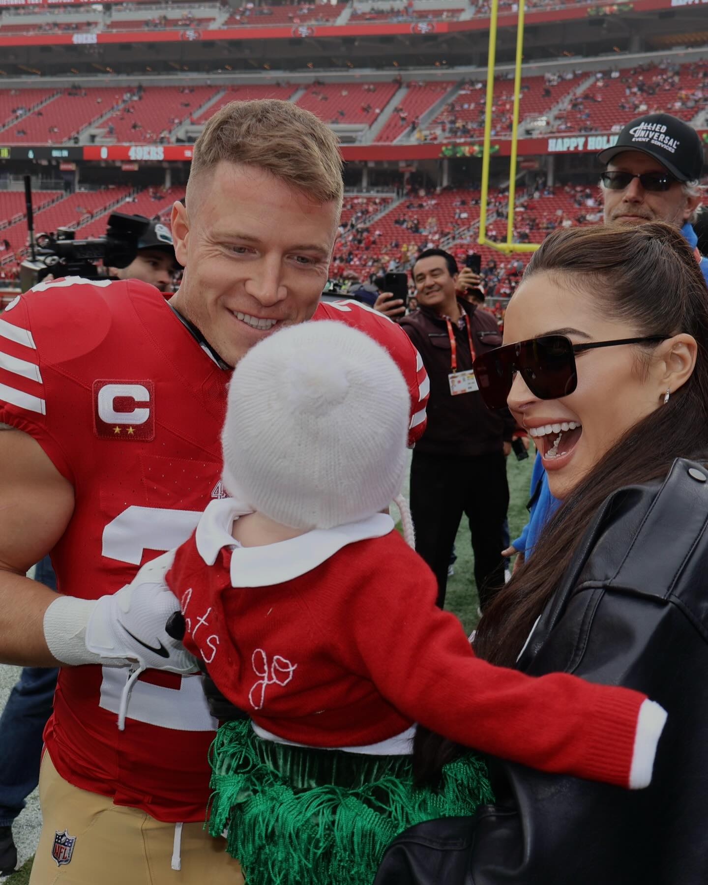 San Francisco 49ers player Christian McCaffrey smiles at his baby, held by Olivia Culpo, on the football field.
