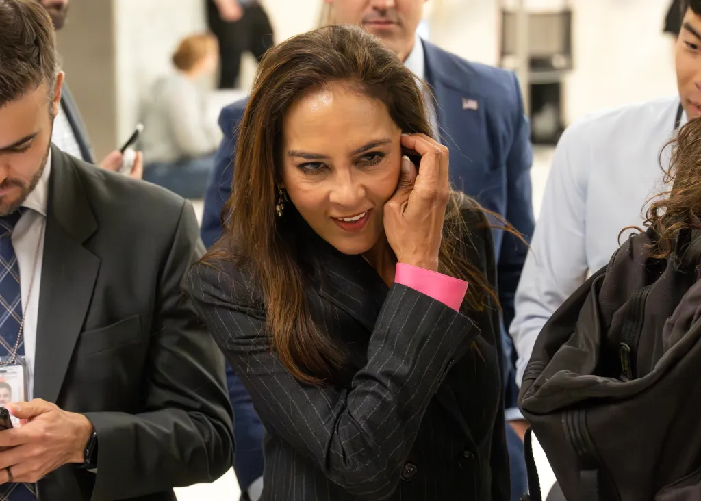 Harmeet K. Dhillon, in a dark pinstriped jacket with a hot pink sleeve, smiling and touching her hair in a crowd.