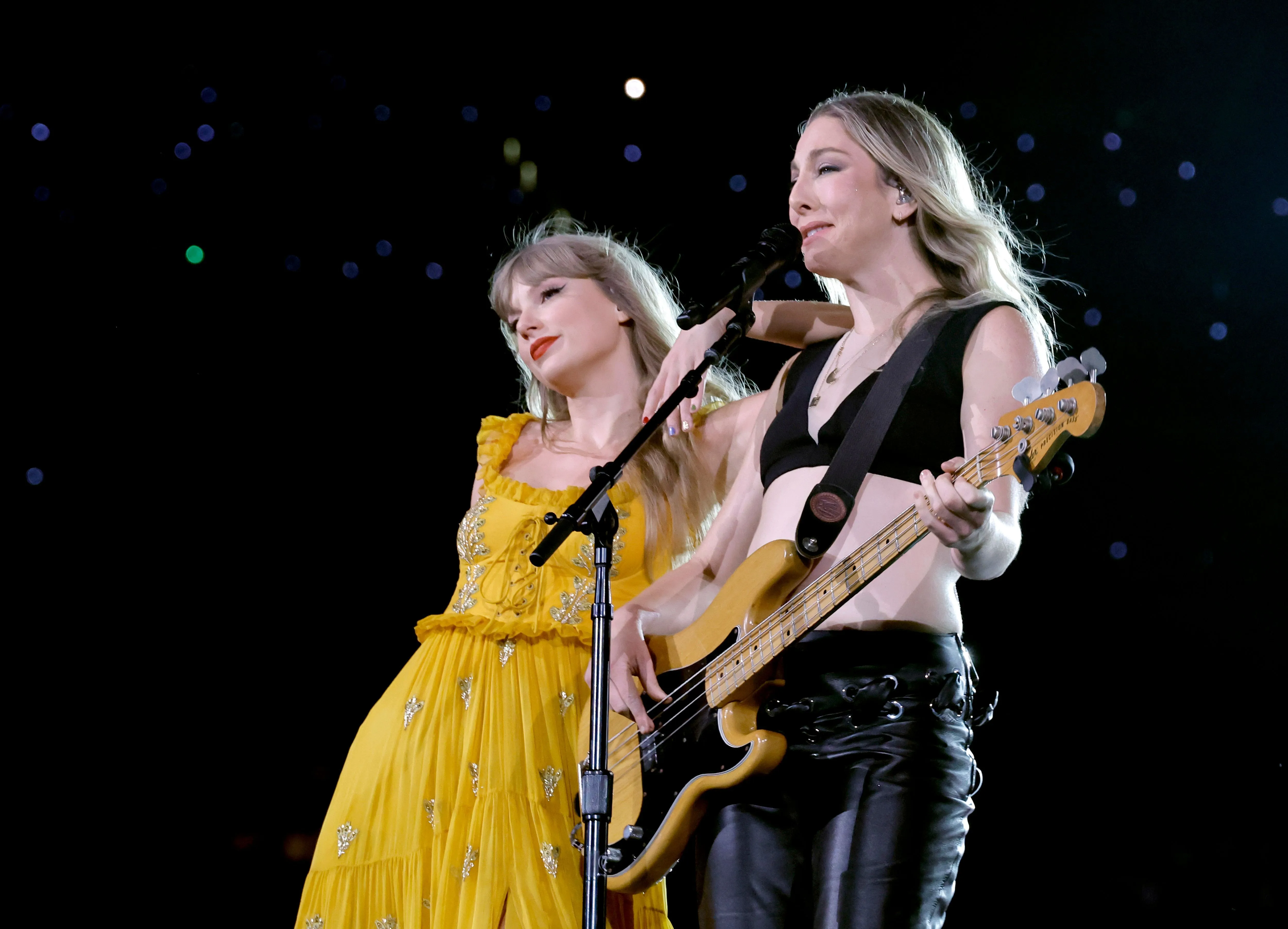 Taylor Swift in a yellow gown and Este Haim of HAIM with a bass guitar perform together on stage.
