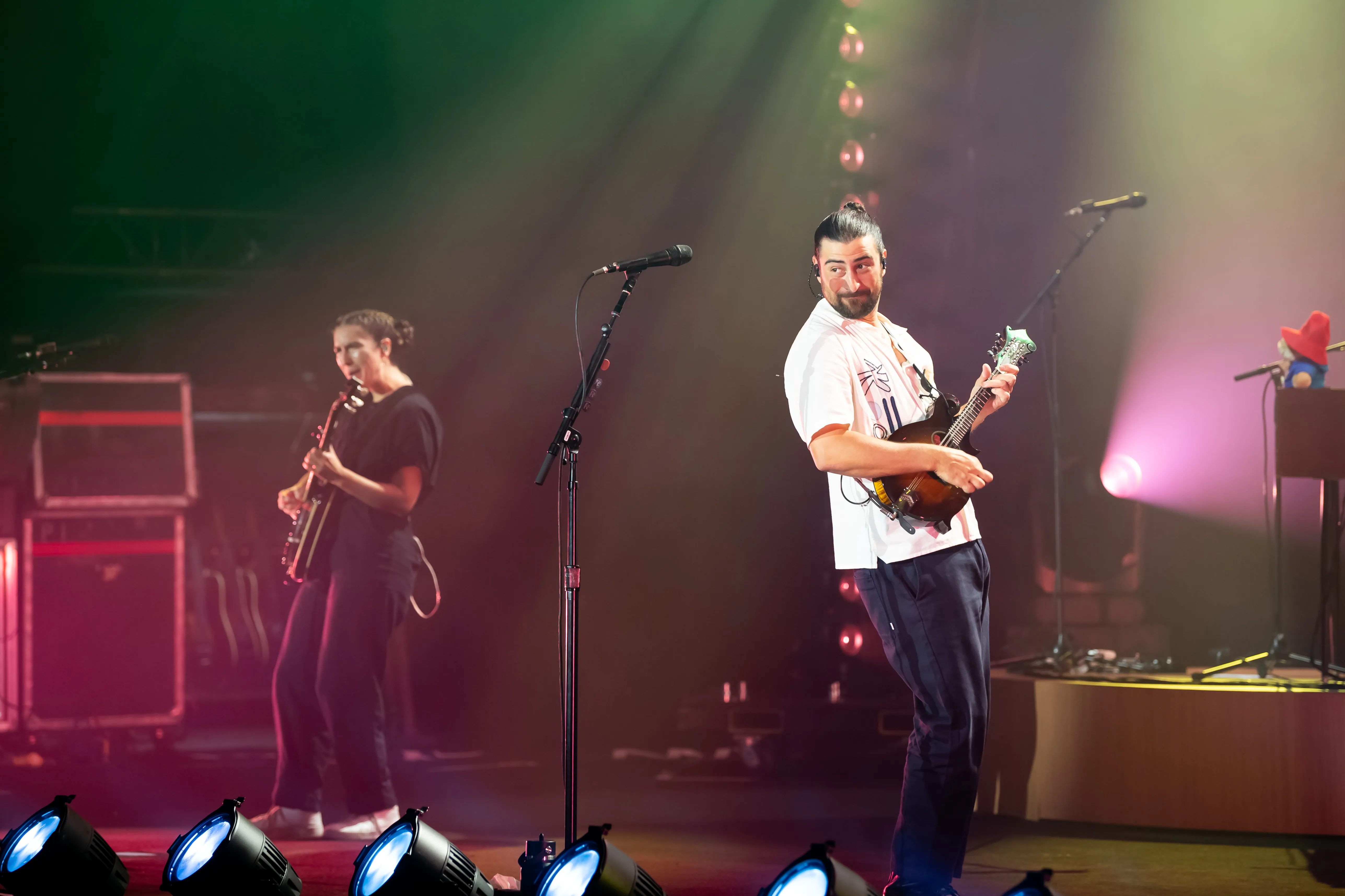 Noah Kahan performing with a mandolin on stage with another musician playing guitar in the background.