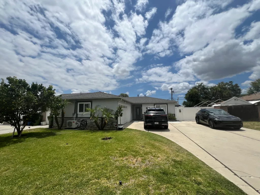 A gray house with two black cars parked in the driveway and a green lawn under a cloudy sky.