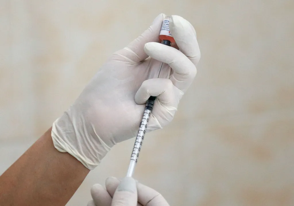 A nurse in white gloves fills a syringe with vaccine from a vial.