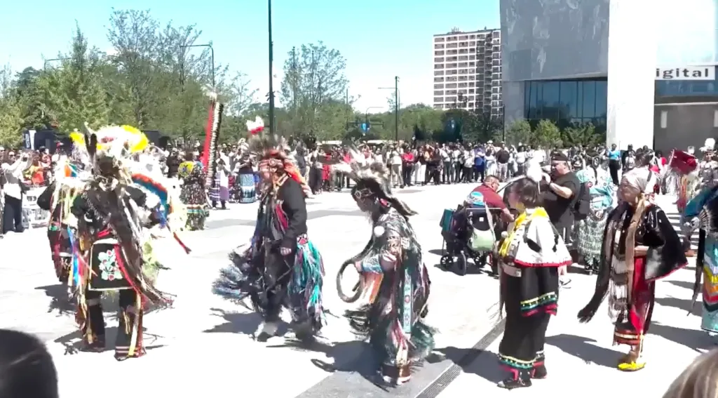 Native American individuals in traditional regalia perform a dance outdoors.