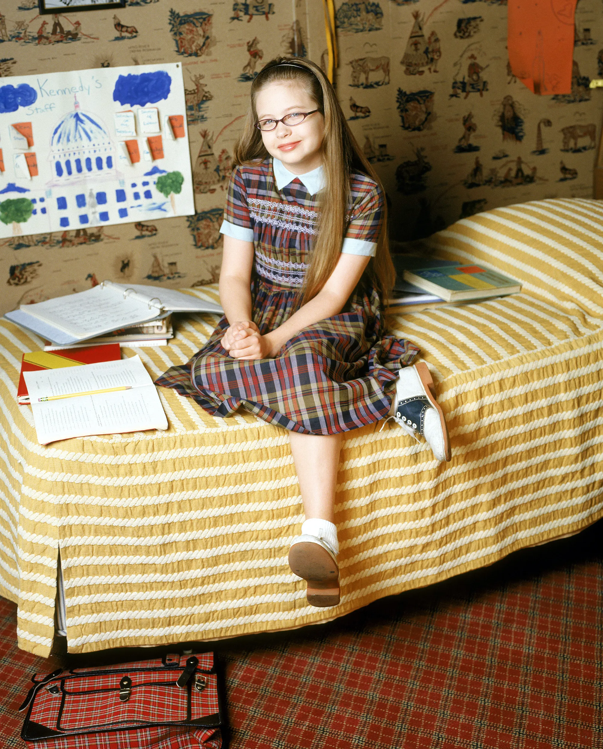 Daveigh Chase studying in a plaid dress and saddle shoes, sitting on a bed in a bedroom.
