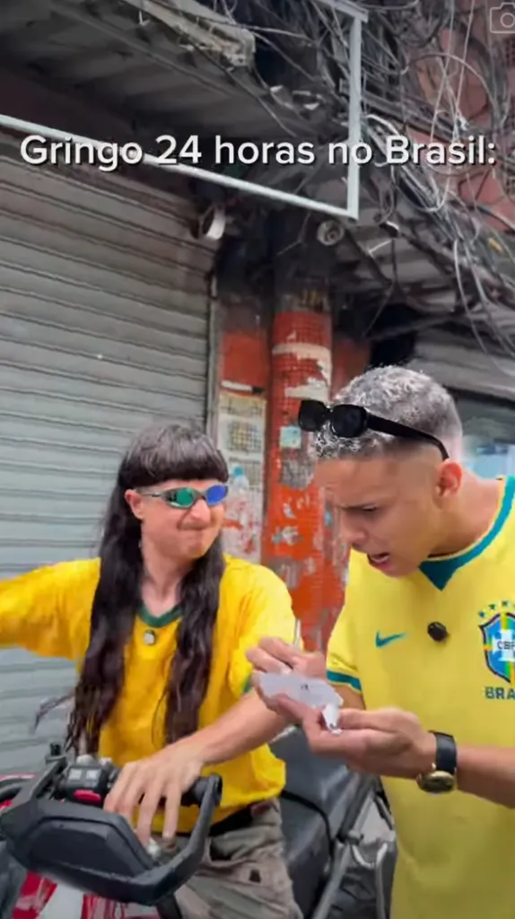 Two men in yellow Brazil jerseys; one with a mullet and sunglasses sits on a motorcycle, the other stands beside him, looking at something in his hand.
