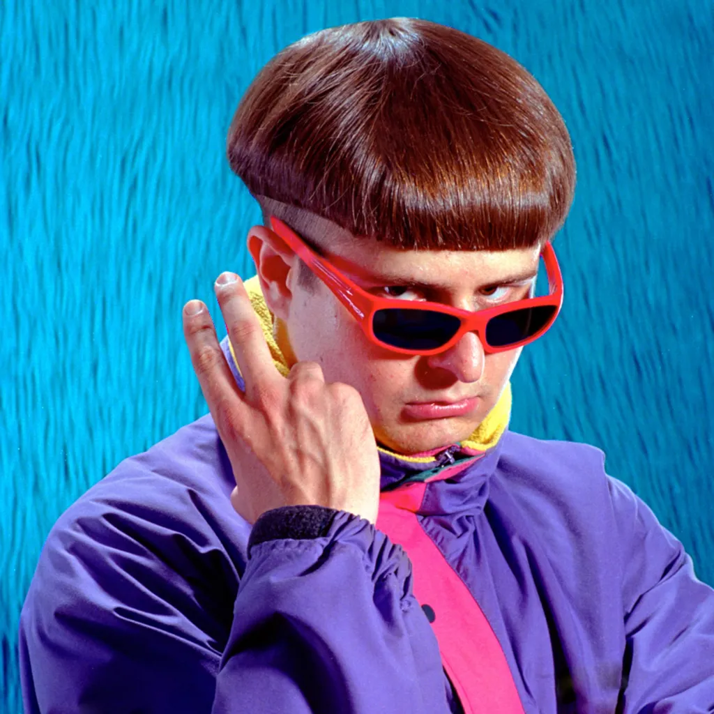 A young man with a bowl cut, red sunglasses, and a purple jacket holds up two fingers.