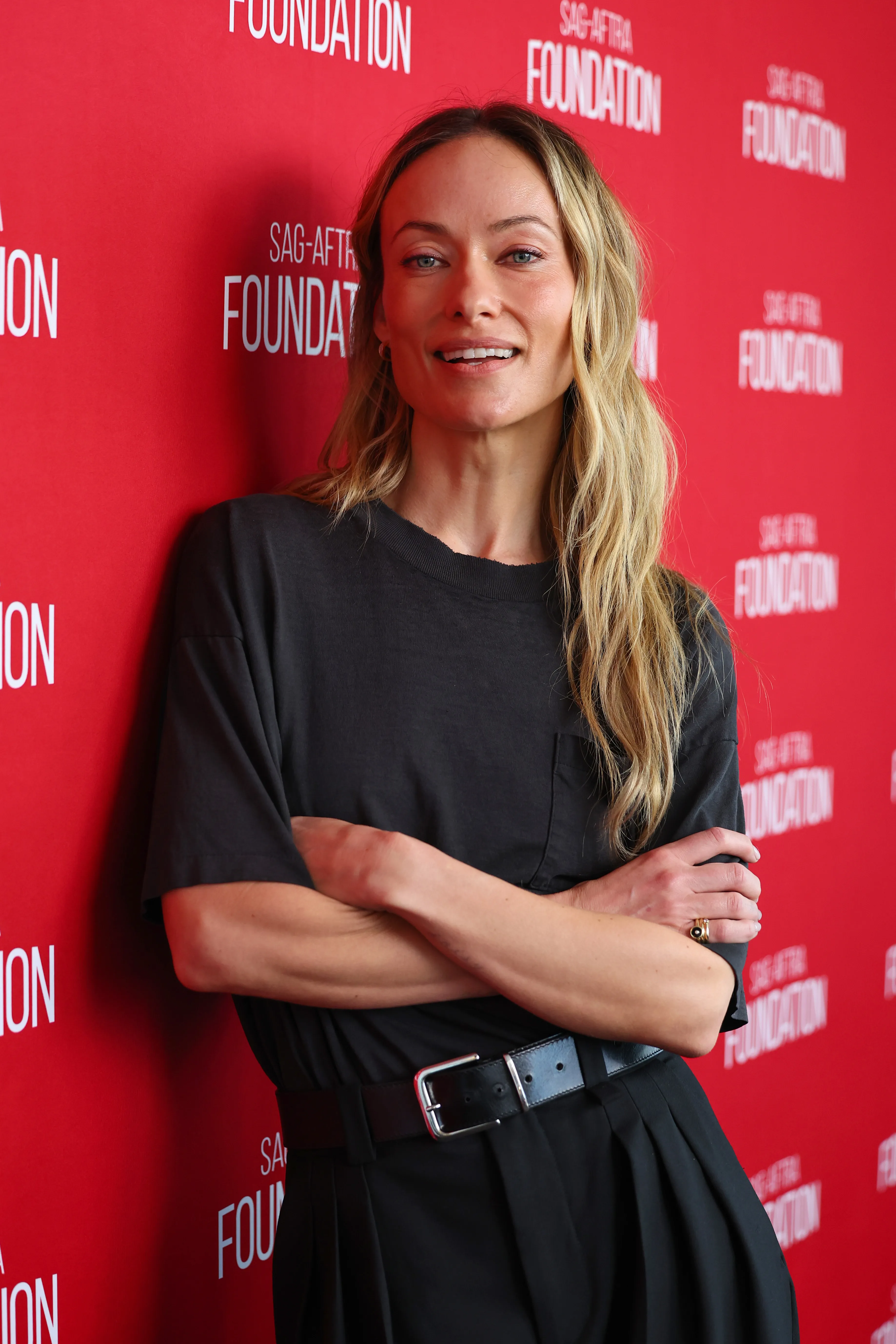 Olivia Wilde smiling with her arms crossed in front of a red backdrop with 