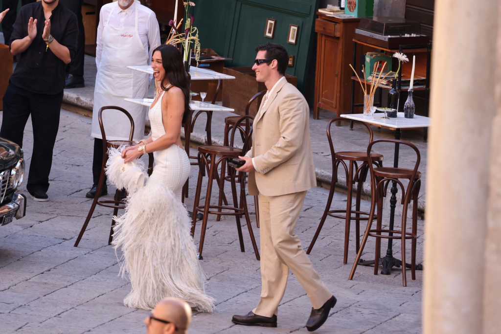 Dua Lipa in a white feathered gown and Callum Turner in a beige suit walk through Palermo.