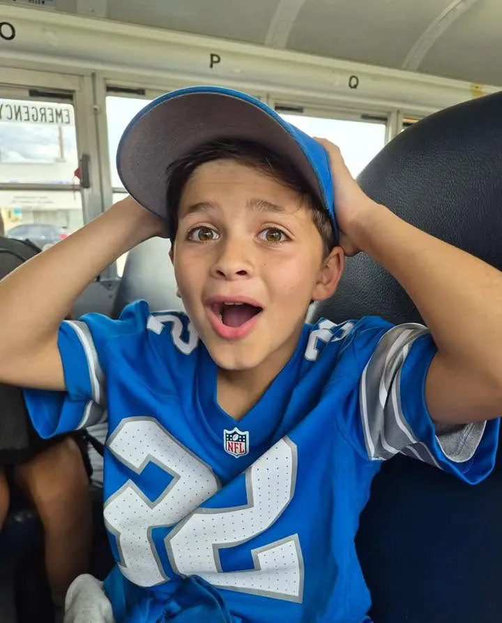 A young boy wearing a blue football jersey and a backwards baseball cap sits with his hands on his head, looking surprised.