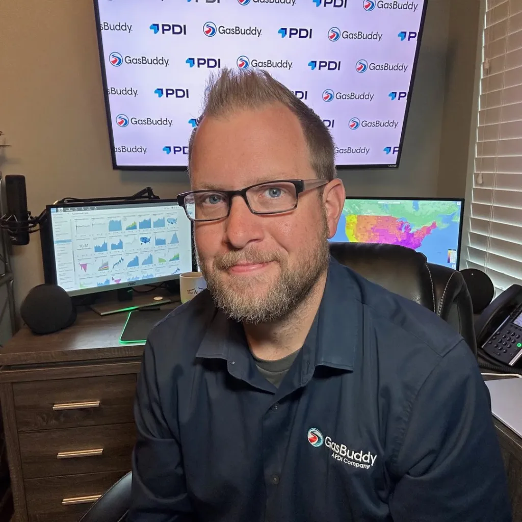Patrick De Haan, wearing a GasBuddy shirt, seated in front of monitors displaying gas price data.