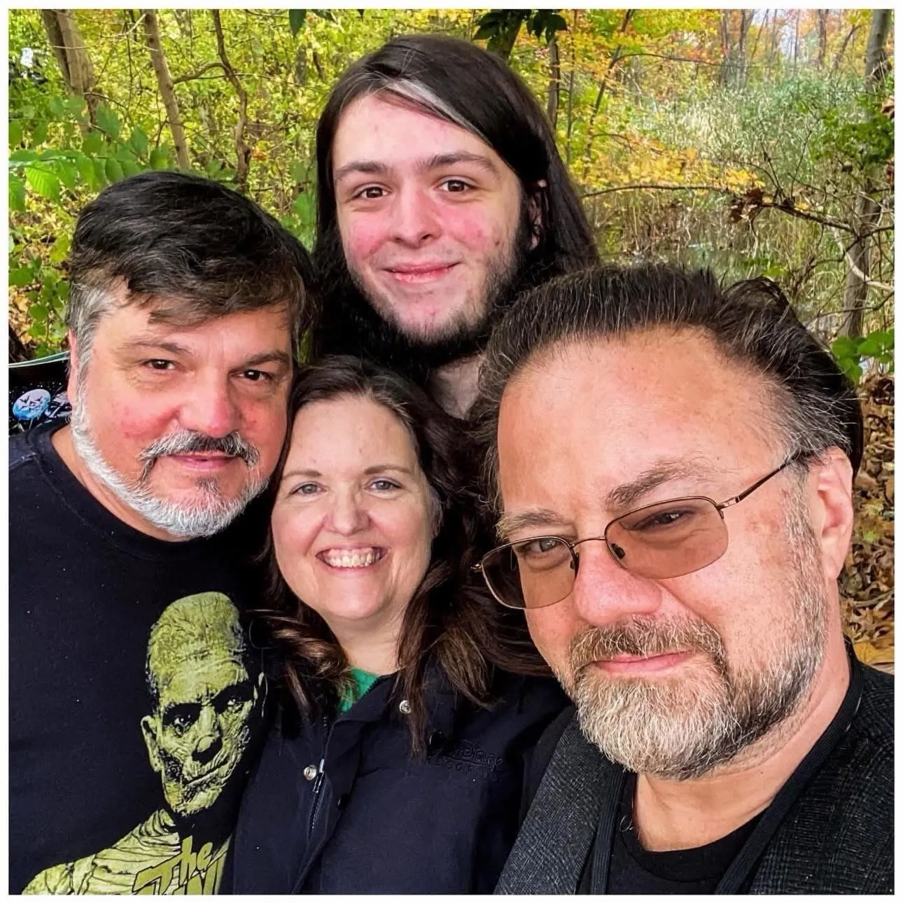 A group of four smiling people, two men, one woman, and one younger man, posing for a picture outdoors.
