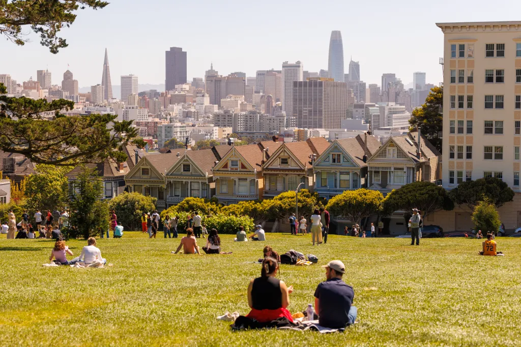 People relax on a grassy hill in Alamo Square Park, with the Painted Ladies Victorian houses and the San Francisco skyline in the background.