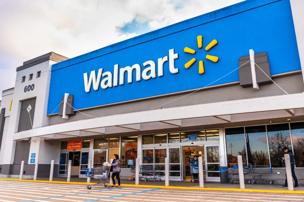 Exterior of a Walmart store with its blue and yellow logo, showing shoppers with carts entering and exiting the entrance.