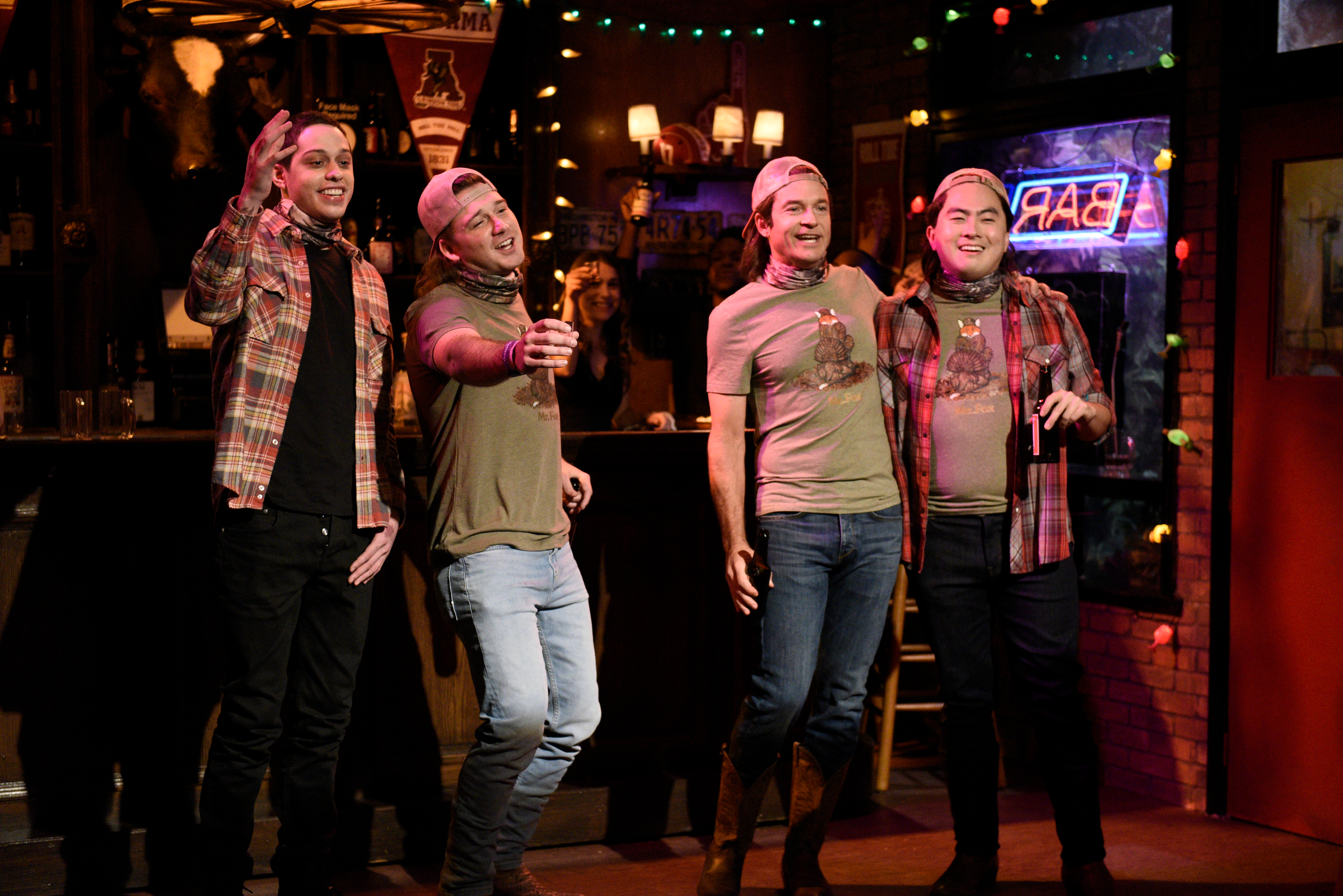 Pete Davidson, Morgan Wallen, Jason Bateman, and Bowen Yang in a comedy sketch, dressed in plaid shirts, neck gaiters, and baseball caps, standing in a bar.