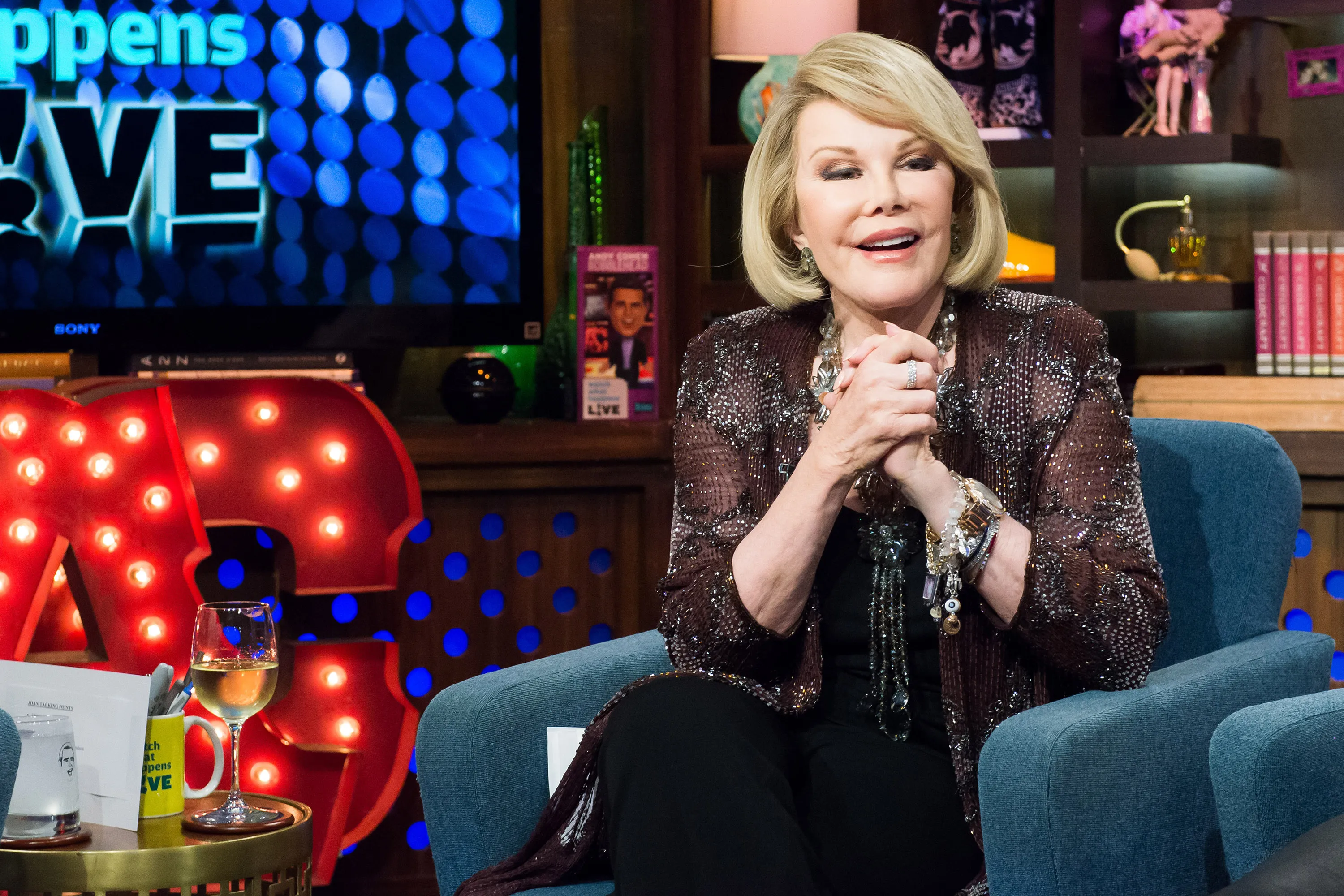 Comedian Joan Rivers seated on a blue armchair, smiling, with a television screen displaying 