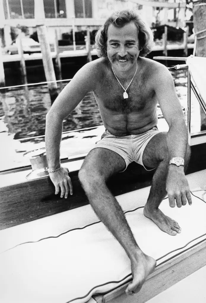 Jimmy Buffett sitting on a boat, smiling, shirtless with a necklace and watch.