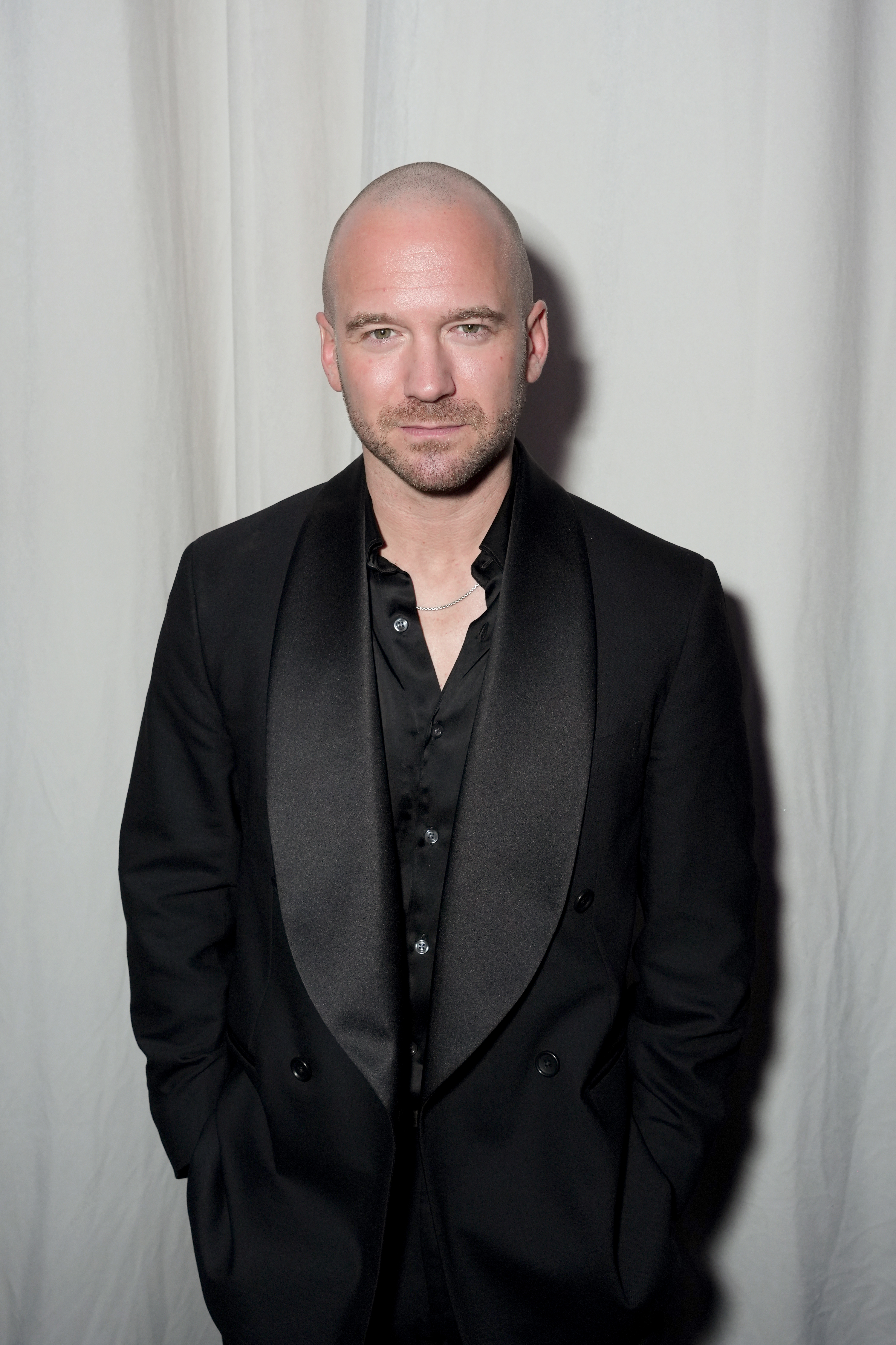 Sean Evans poses in a black suit and black collared shirt with a silver chain against a white backdrop.