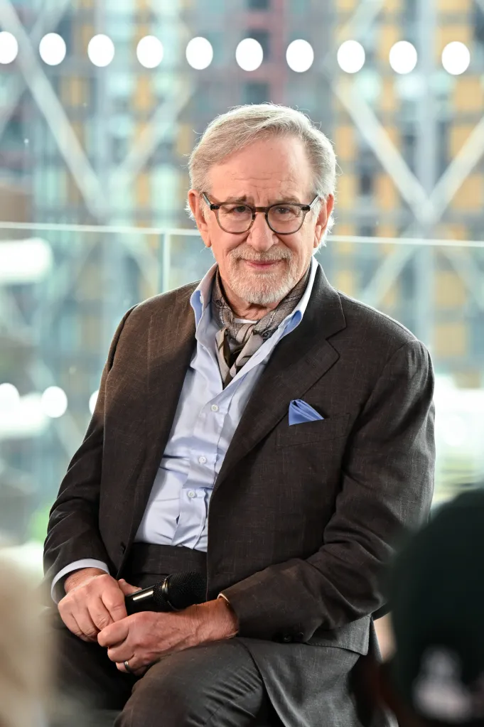 Steven Spielberg in a gray suit and light blue shirt, holding a microphone.