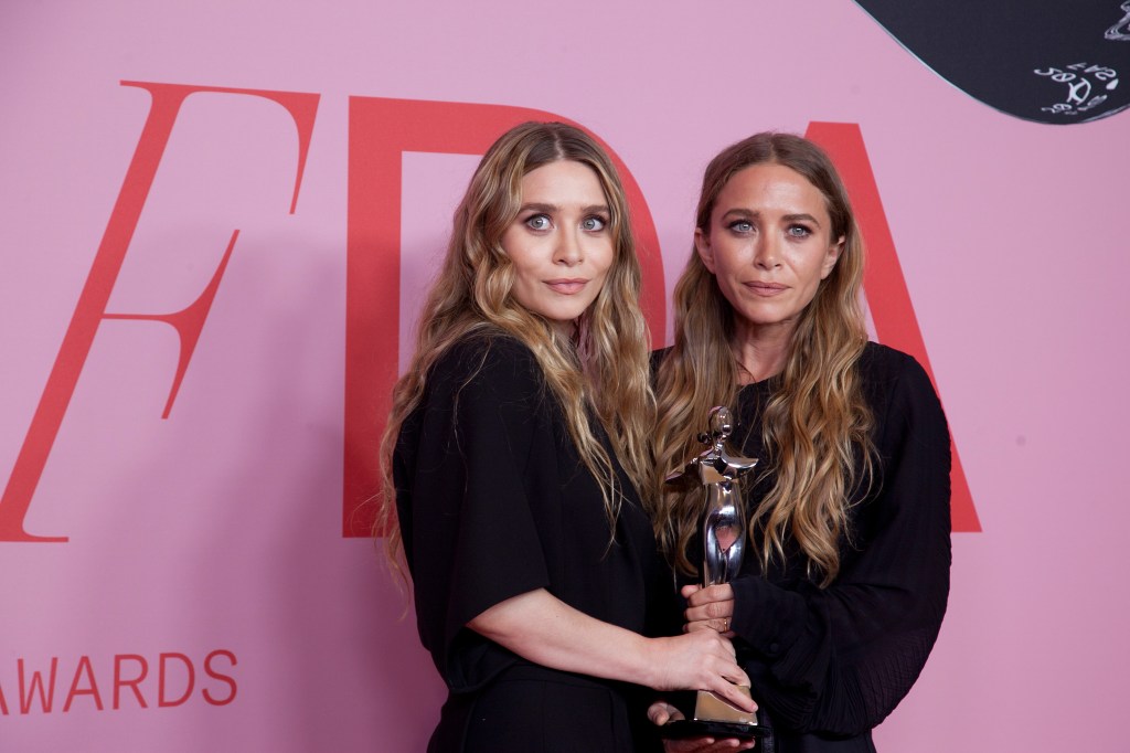Ashley and Mary-Kate Olsen holding a silver trophy in front of a pink and red CFDA Fashion Awards backdrop.