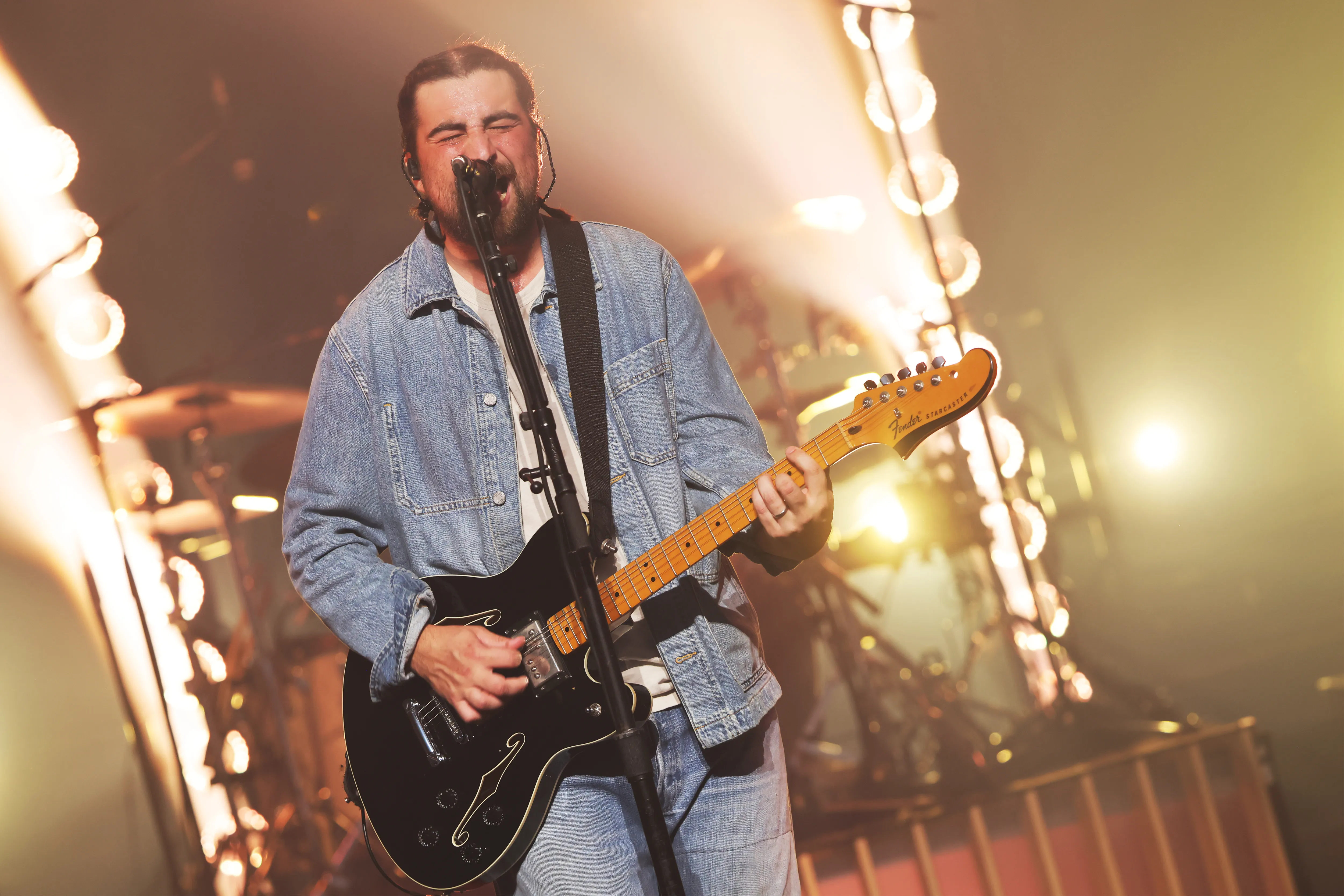 Noah Kahan performing live, playing a black electric guitar and singing into a microphone.