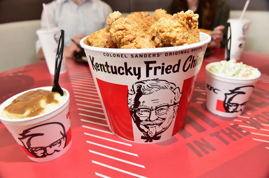 KFC bucket of fried chicken, mashed potatoes, and coleslaw on a red table.