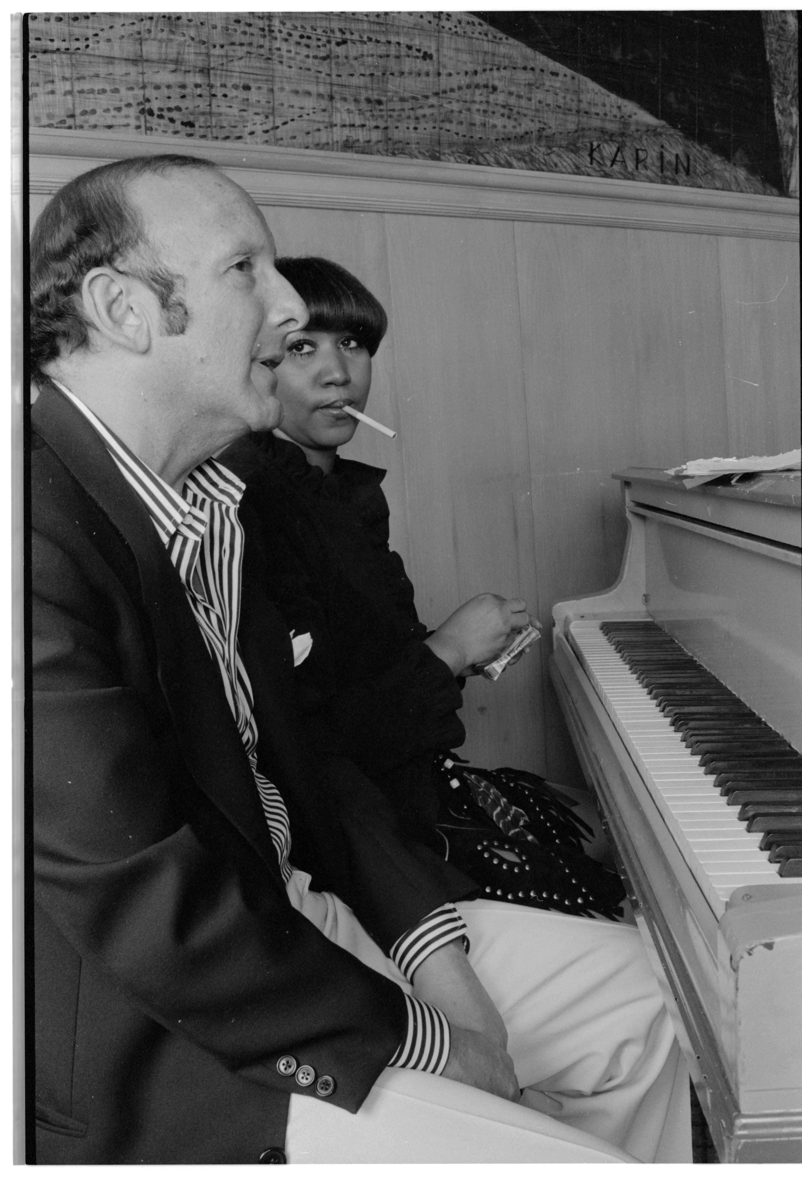 Aretha Franklin smoking a cigarette, sitting next to Clive Davis at a piano.