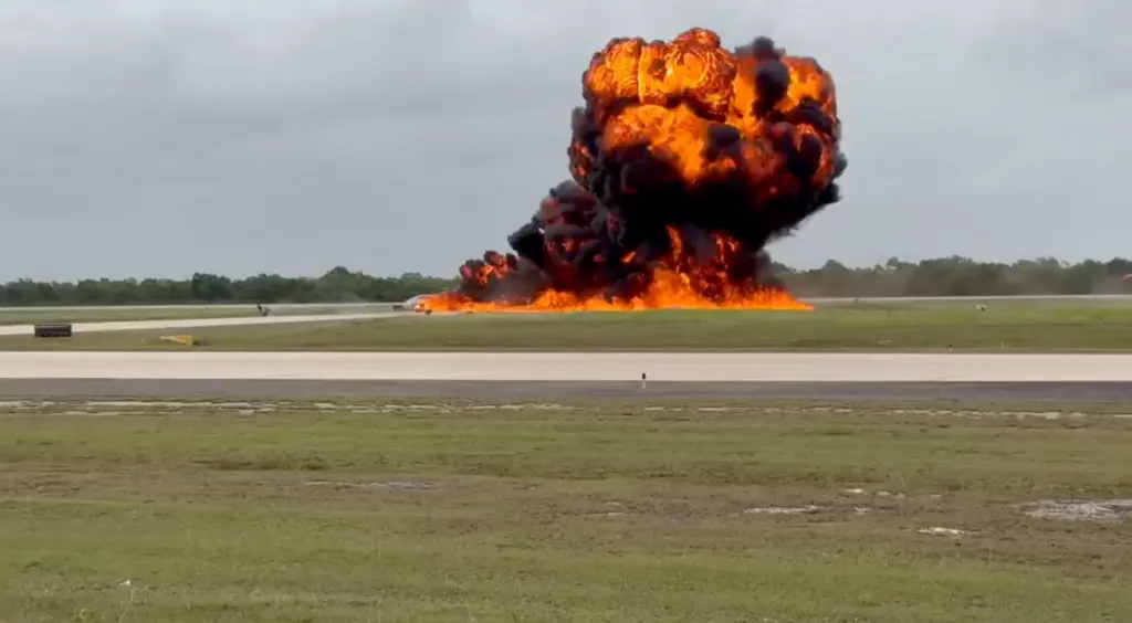 A massive fire and black smoke plume from a crashed private jet on a runway.